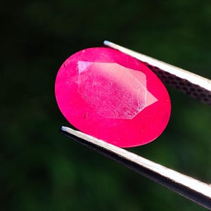 Könnte beinhalten: Ein oval geschliffener Rubin, gehalten von einer silbernen Pinzette. Der Rubin hat eine leuchtende, tiefrosa Farbe mit einer reflektierenden Oberfläche. Der Hintergrund ist unscharf.