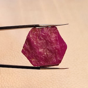 May include: A deep pink, faceted ruby gemstone held by tweezers. The ruby has a rough, natural appearance.