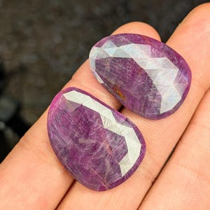 May include: Two faceted, oval-shaped ruby gemstones. The gems are a deep magenta color with white, geometric facets. The stones are displayed on a neutral background.