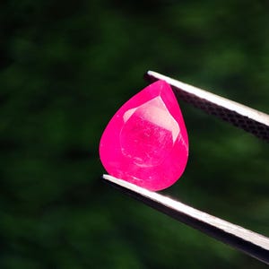 Può includere: Una gemma rosa vibrante a forma di pera sfaccettata è tenuta da una pinzetta. La gemma ha una superficie lucida e un colore brillante e saturo. Lo sfondo è un verde scuro sfocato.