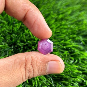 Puede incluir: Una gema hexagonal púrpura facetada, sostenida entre dos dedos. La gema tiene un patrón interno complejo y una superficie pulida, que refleja la luz. El fondo es verde borroso, lo que sugiere un entorno natural. El color de la gema varía de púrpura claro a oscuro.