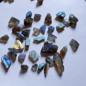 Puede incluir: Una colección de virutas de piedra preciosa de labradorita iridiscente en varios tonos de azul, verde y dorado. Las virutas tienen una forma irregular y una textura natural y rugosa.