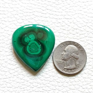 May include: A green malachite gemstone in the shape of a guitar pick, next to a US quarter for size comparison.