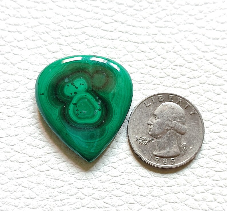 May include: A green malachite gemstone in the shape of a guitar pick, next to a US quarter for size comparison.