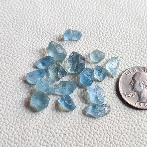 Pu&ograve; includere: Una collezione di piccole pietre preziose di acquamarina blu chiaro. Le pietre preziose sono grezze e hanno una variet&agrave; di forme e dimensioni. Le pietre preziose sono disposte su una superficie bianca accanto a una moneta da un euro per la scala.