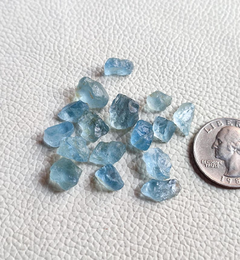 Pu&ograve; includere: Una collezione di piccole pietre preziose di acquamarina blu chiaro. Le pietre preziose sono grezze e hanno una variet&agrave; di forme e dimensioni. Le pietre preziose sono disposte su una superficie bianca accanto a una moneta da un euro per la scala.