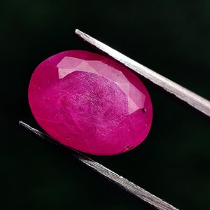 Può includere: Una gemma di rubino sfaccettata, di forma ovale, tenuta da pinzette metalliche su uno sfondo scuro. Il rubino mostra un colore rosso intenso e vibrante con texture interne e riflessi di luce.