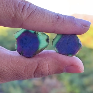 May include: Two raw gemstones are held between fingers. Each stone has a dark blue centre, surrounded by a ring of purple and a green outer edge. The stones have a rough, natural texture.