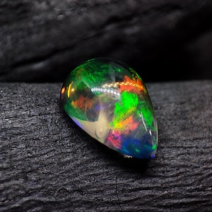 Peut inclure: Une pierre d'opale polie en forme de goutte d'eau. L'opale présente un jeu de couleurs vibrant, avec des éclairs de vert, de rouge, d'orange et de bleu. La pierre est posée sur un fond sombre et texturé, soulignant ses qualités iridescentes.