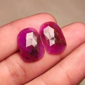 May include: Two faceted oval-shaped gemstones with a pink and purple hue. The gemstones have a faceted cut and are set in a bezel setting.