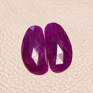 Może przedstawiać: Dwa fioletowe kaboszony z kamieni szlachetnych o fasetowanym wzorze. Kamienie mają owalny kształt i błyszczące wykończenie.
