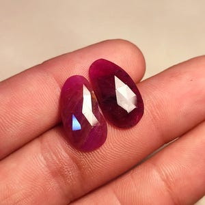 May include: Two faceted, oval-shaped, deep red gemstones. The gems have a polished, reflective surface with multiple facets that catch the light, creating bright highlights. The gems are displayed on a neutral background.