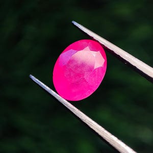 Puede incluir: Una gema rosa intenso, facetada y de forma ovalada, es sostenida por unas pinzas plateadas sobre un fondo verde borroso. El tallado de la gema crea múltiples superficies reflectantes, realzando su color vibrante. Las pinzas proporcionan un agarre seguro, mostrando las facetas pulidas.