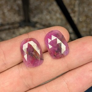 May include: Two oval-shaped, faceted, pink gemstones. The gems have a mix of light and dark pink hues with internal inclusions. The facets create a geometric pattern, reflecting light. The gemstones are displayed against a neutral background.