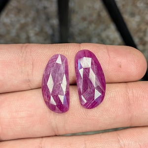 May include: Two faceted, oval-shaped, deep magenta gemstones. The gems have a polished surface with a geometric pattern of reflective facets. The stones are held in a hand, showcasing their color and cut. These gemstones are suitable for jewelry making.
