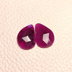 May include: Two faceted, teardrop-shaped gemstones with a deep purple hue. The stones have a faceted, diamond-like cut.