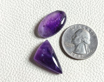 May include: Two amethyst gemstones, one oval shaped and one triangular, on a white textured surface. A US quarter is included for size reference.