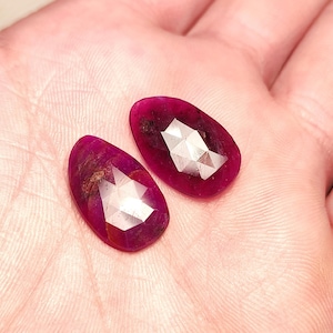 Puede incluir: Dos piedras preciosas de rubí facetadas con un respaldo plateado. Las piedras preciosas son de forma ovalada y tienen un color rojo intenso.