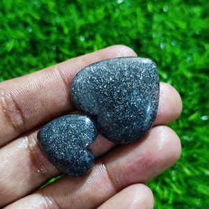 Peut inclure: Deux pierres en forme de cœur avec une finition gris foncé scintillante. Les pierres sont tenues dans une main.