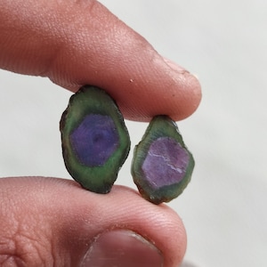 Może przedstawiać: Dwa surowo cięte kamienie szlachetne z turmalinu arbuzowego. Kamienie mają zieloną warstwę zewnętrzną, fioletowy środek i jasne wnętrze. Kamienie szlachetne są trzymane między palcami na białym tle.