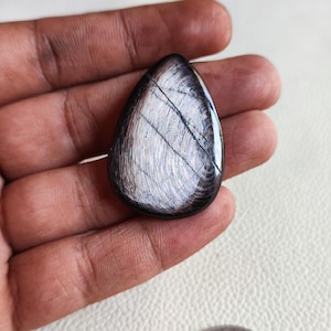 May include: A teardrop-shaped cabochon gemstone with a black and white iridescent finish. The stone has a smooth, polished surface and a unique pattern of lines.