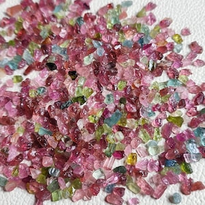 May include: A large collection of small, colourful, rough-cut gemstones. The gemstones are a mix of pink, green, blue, and clear colours. They are scattered on a white surface.