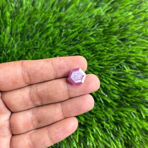 Peut inclure: Une pierre précieuse rose à facettes avec un motif géométrique. La gemme est tenue dans une main, sur un fond d'herbe artificielle verte. Les facettes de la gemme reflètent la lumière, créant un effet scintillant.