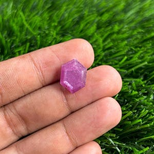 Puede incluir: Una gema rosa facetada de forma hexagonal. La gema se sostiene en una mano sobre un fondo de hierba verde. Las facetas de la gema capturan la luz, creando un efecto brillante. Esta gema es un hermoso ejemplo de formaciones minerales naturales.