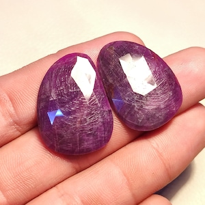 May include: Two purple gemstone cabochons with a faceted cut. The stones are oval shaped and have a slightly irregular shape. The stones are translucent and have a slight shimmer.