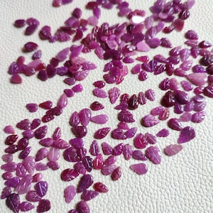 May include: A large collection of loose purple gemstone leaf shapes. The gemstones are scattered on a white surface.