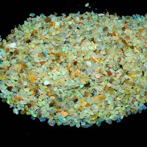 Op de afbeelding: Een grote hoop kleine, iriserende opaalchips in verschillende tinten groen, blauw en geel. De chips zijn verspreid over een zwarte achtergrond.