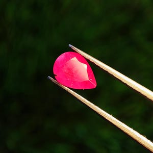 Könnte beinhalten: Ein facettierter, tropfenförmiger Rubin-Edelstein, der von einer Pinzette gehalten wird. Der Rubin hat eine leuchtende, tiefrote Farbe und reflektiert das Licht. Der Hintergrund ist ein verschwommenes, dunkles Grün.
