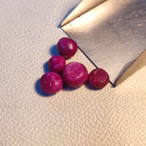 Może przedstawiać: Pięć luźnych, okrągłych, różowych rubinów na białej, teksturowanej powierzchni. Kamienie szlachetne są różnych rozmiarów i mają gładkie, wypolerowane wykończenie.