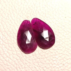 Może przedstawiać: Dwa fasetowane, gruszkowate kamienie szlachetne w głębokim odcieniu różowym. Kamienie są oszlifowane fasetowaną formą i mają przezroczysty wygląd.