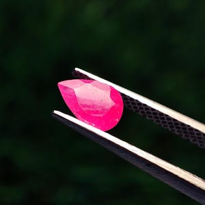Op de afbeelding: Een gefacetteerde, druppelvormige robijn, vastgehouden door een pincet. De robijn heeft een levendige, dieproze kleur, met zichtbare facetten die het licht vangen. De achtergrond is een vage, donkergroene kleur.
