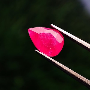 Op de afbeelding: Een gefacetteerde, druppelvormige robijn edelsteen, vastgehouden door een pincet. De robijn heeft een levendige, dieproze kleur, met meerdere facetten die het licht vangen. De achtergrond is een vage, donkergroene kleur.