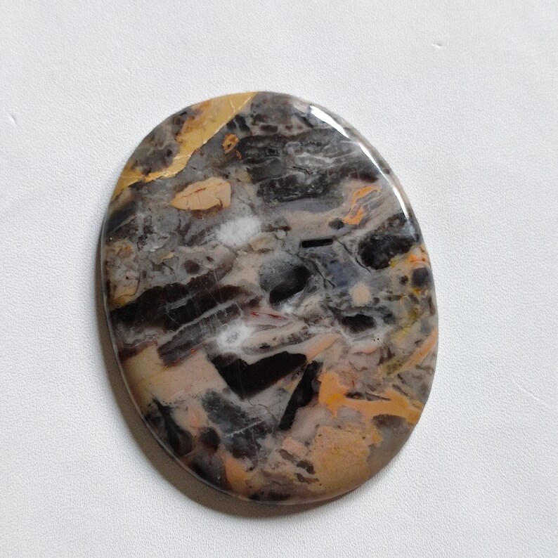 May include: A round, oval-shaped cabochon gemstone with a swirling pattern of black, brown, white, and gold. The stone has a polished, smooth surface.