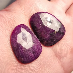 May include: Two purple faceted gemstones with a white geometric design. The stones are oval shaped and have a smooth, polished surface.