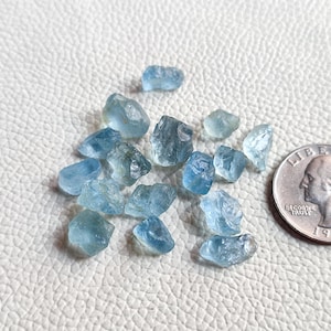 Pu&ograve; includere: Una collezione di piccole pietre preziose di acquamarina ruvide di colore blu chiaro. Le pietre sono sparse su una superficie bianca accanto a una moneta da 25 centesimi di dollaro americano per la scala.