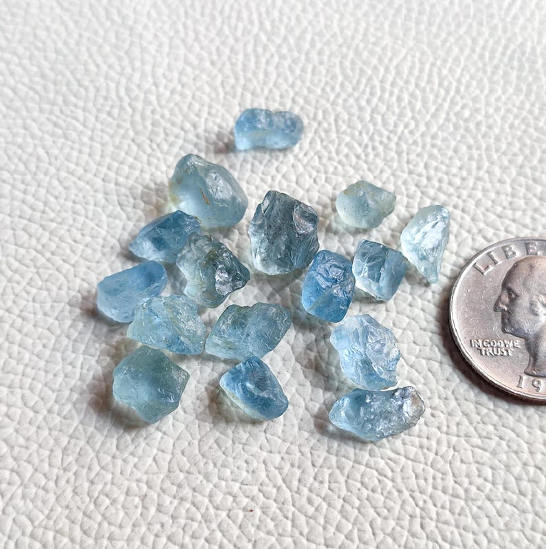 Pu&ograve; includere: Una collezione di piccole pietre preziose di acquamarina ruvide di colore blu chiaro. Le pietre sono sparse su una superficie bianca accanto a una moneta da 25 centesimi di dollaro americano per la scala.