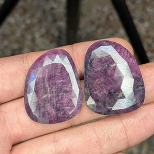 May include: Two faceted, oval-shaped, deep purple gemstones. The gems have a polished, reflective surface with internal veining. The stones are held in a hand, showcasing their color and cut. The gems are likely rubies or similar precious stones.
