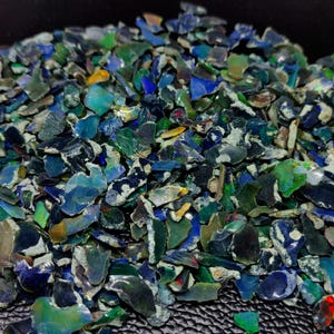 Puede incluir: Un montón de virutas de ópalo iridiscentes en tonos azules, verdes y negros. Los fragmentos tienen un aspecto rugoso y fracturado, con destellos de color visibles. Los ópalos probablemente se utilizan para joyería o decoración.