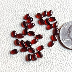 Peut inclure: Une collection d'environ 25 pierres précieuses ovales de couleur rouge foncé, probablement des grenats, éparpillées sur une surface blanche. Un quart de dollar américain est inclus pour l'échelle.