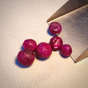 May include: Six faceted ruby gemstones in various sizes and shapes, displayed on a white surface with a metal scoop.
