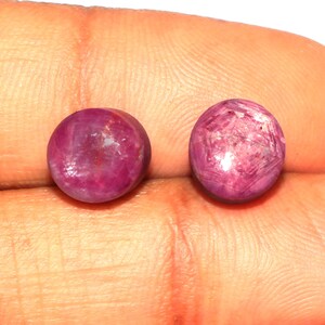 Könnte beinhalten: Zwei runde, rosafarbene Rubingesteine mit einer glatten, polierten Oberfläche. Die Edelsteine sind in einer Zargenfassung gefasst und haben einen leichten Glanz.
