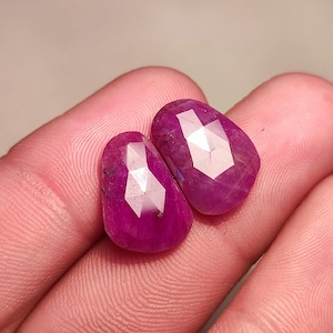 Może przedstawiać: Dwa fasetowane różowe rubiny, każdy o unikalnym kształcie i szlifie, są prezentowane na palcu.