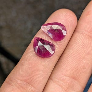 May include: Two faceted, teardrop-shaped, deep red gemstones. The gems are held in a hand, with the facets catching the light. The stones have a polished, reflective surface, showcasing their rich color and clarity. These gems are likely rubies.