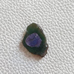 May include: A raw gemstone with a dark green outer layer and a central, circular area of deep purple. The stone has a natural, irregular shape and texture, suggesting it is unpolished.
