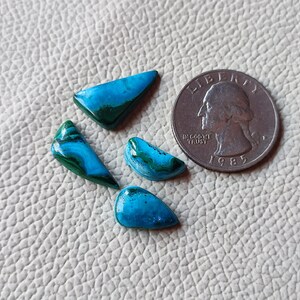 Puede incluir: Cuatro cabujones de piedras preciosas turquesa y verdes de formas y tamaños variados, incluyendo un triángulo, una media luna y una lágrima, están dispuestos sobre una superficie blanca texturizada. Una moneda de veinticinco centavos de dólar estadounidense de plata se incluye para la escala.