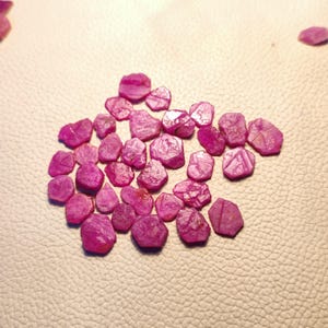 May include: A collection of pink ruby gemstones, each with a unique shape and facet. The rubies are arranged in a loose cluster on a white surface.